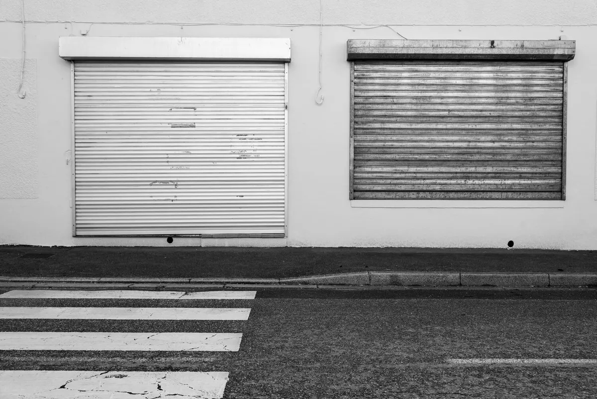 Deux rideaux métalliques à lames baissés devant des devantures, au bord d'un trottoir et d'un passage piéton, photo en noir et blanc