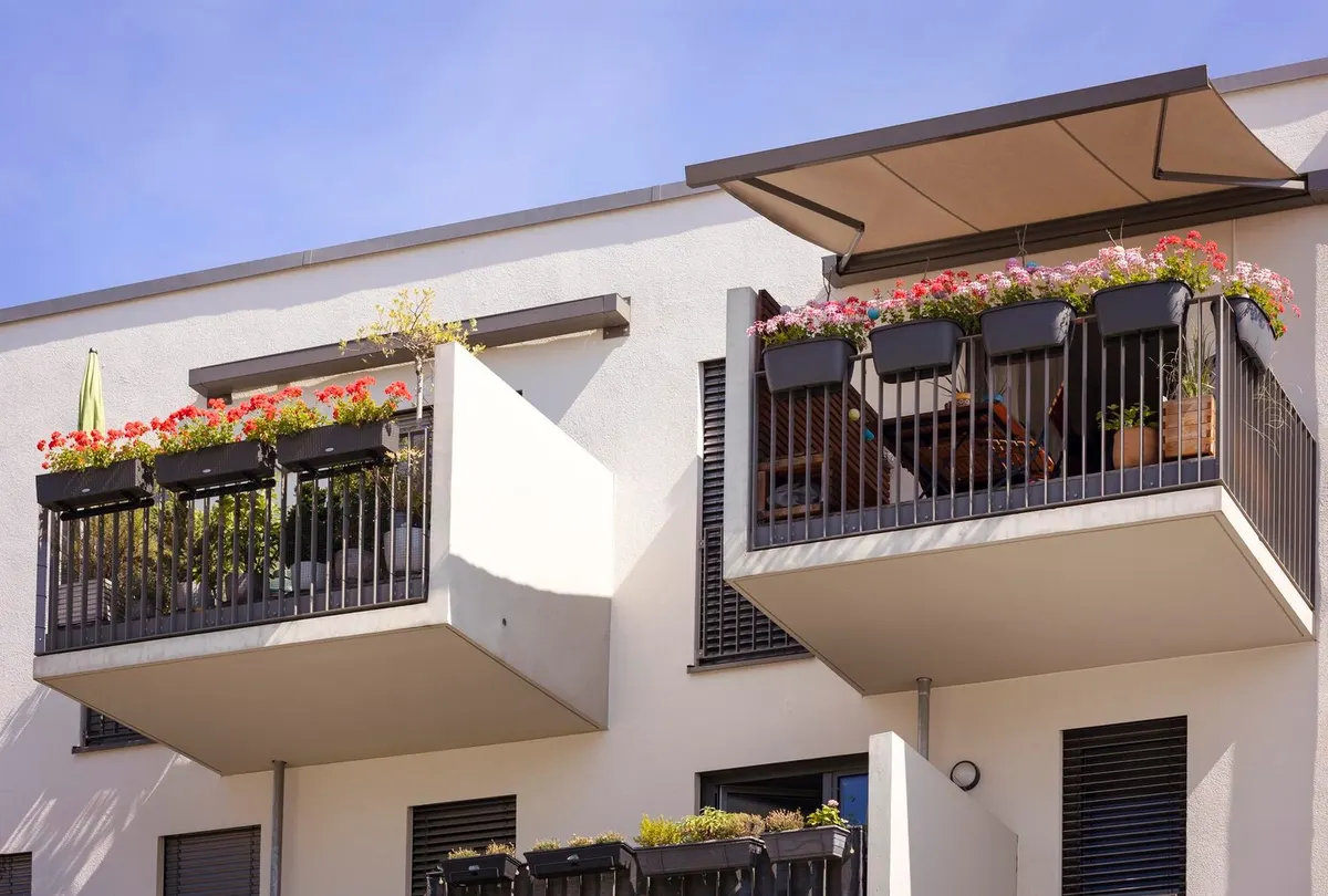 Balcons fleuris d'un immeuble récent à façade blanche, l'un abrité par un store banne à toile beige déployé, fenêtres du dessous fermées par des volets roulants à lames