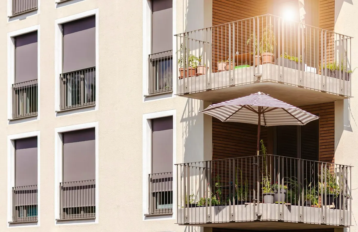 Façade d'immeuble collectif récent aux fenêtres équipées de volets roulants gris presque baissés, à côté de balcons à garde-corps métalliques garnis de plantes